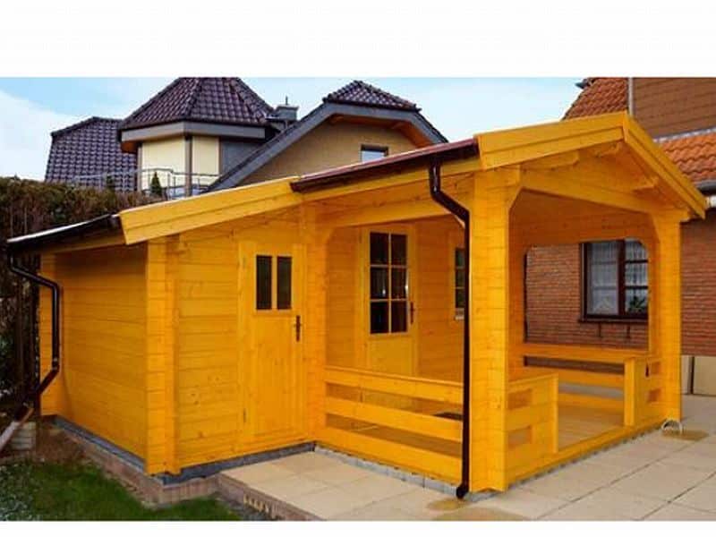 A small bright yellow wooden cabin with a porch, situated in a residential area with larger houses in the background, under a clear sky, serving as a log house model of Carinthia.