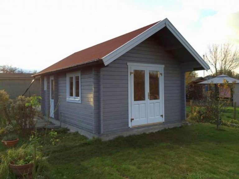 Ein kleines, einstöckiges graues Haus mit rotem Dach und weißen Doppeltüren, das in der Abenddämmerung in einem grasbewachsenen Hof steht.