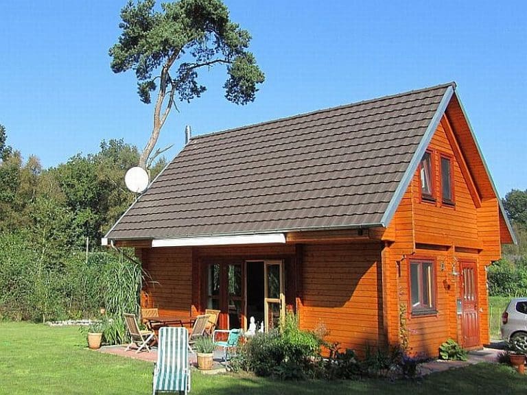 Een klein, oranje houten huisje met een steil bruin dak, gelegen in een tuin met weelderig groen gras en diverse planten. een unieke hoge boom buigt dramatisch over het huis tegen een helderblauwe lucht.