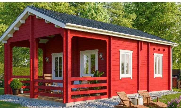 Ein kleines rotes Blockhaus Berlin als Ferienhaus mit weißer Zierleiste und einer überdachten Veranda steht auf einem Kiesgrundstück, umgeben von grünen Bäumen und Pflanzen. Auf der Veranda sind Stühle und ein Tisch aufgestellt.