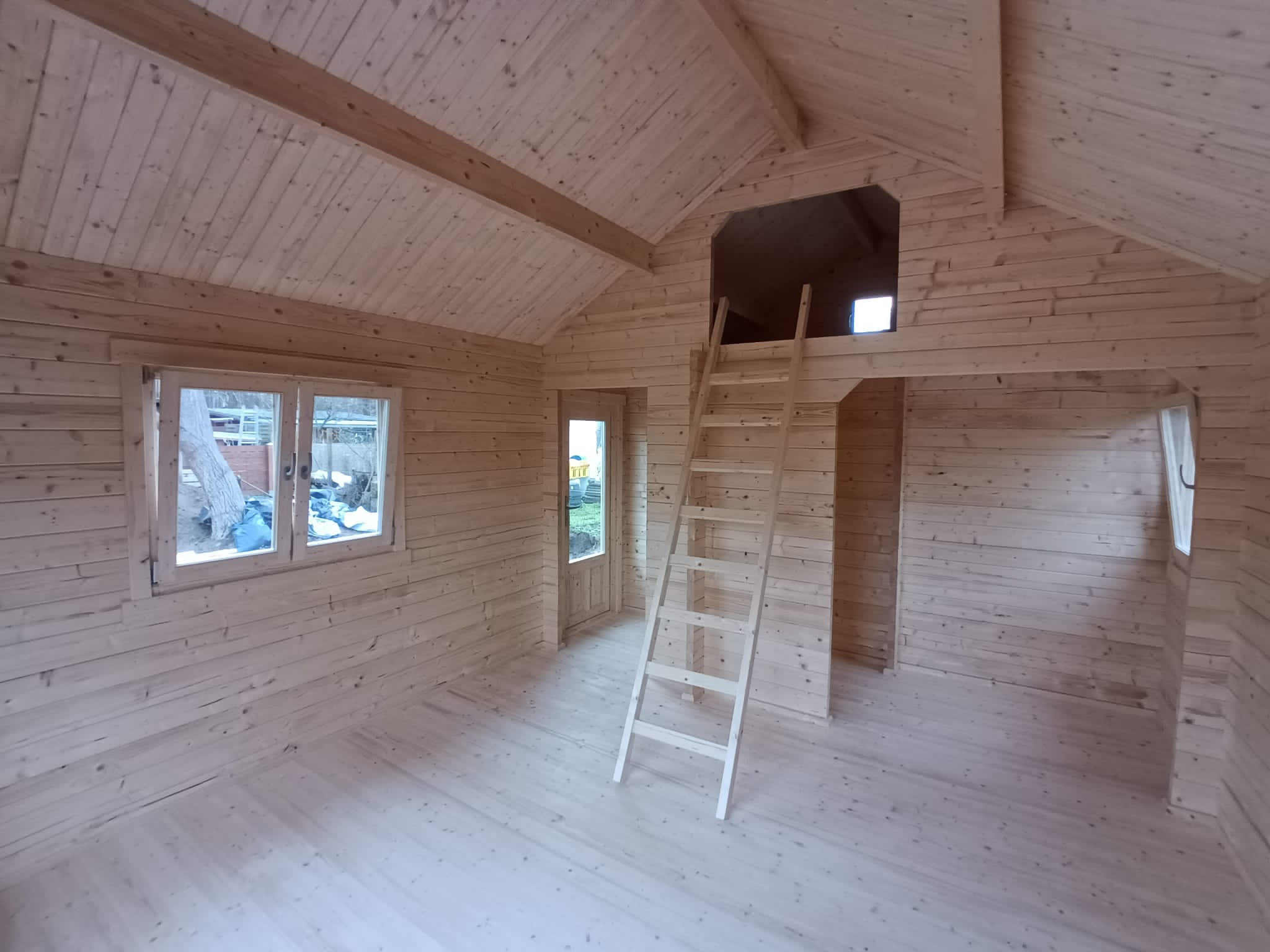 Interieur van een kleine, ongemeubileerde houten hut met zichtbare balken, grote ramen en een zolder bereikbaar via een ladder. de ruimte is leeg en helder, met natuurlijk licht dat naar binnen filtert.