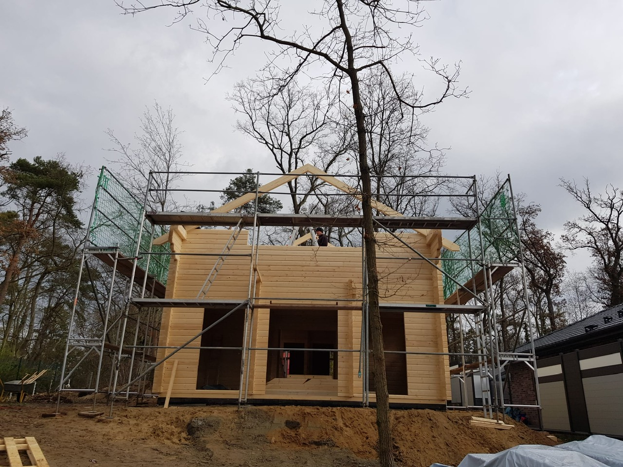 Ein im Bau befindliches zweistöckiges Holzblockhaus mit Gerüst darum und einem Arbeiter auf dem Dach, vor dem Hintergrund kahler Bäume und eines bewölkten Himmels.