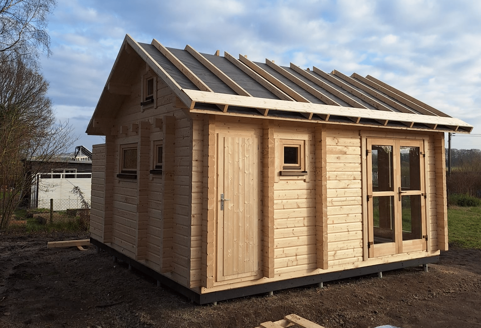 Ein kleines, im Bau befindliches Holzblockhaus mit unvollständiger Überdachung und klarem Himmel im Hintergrund.