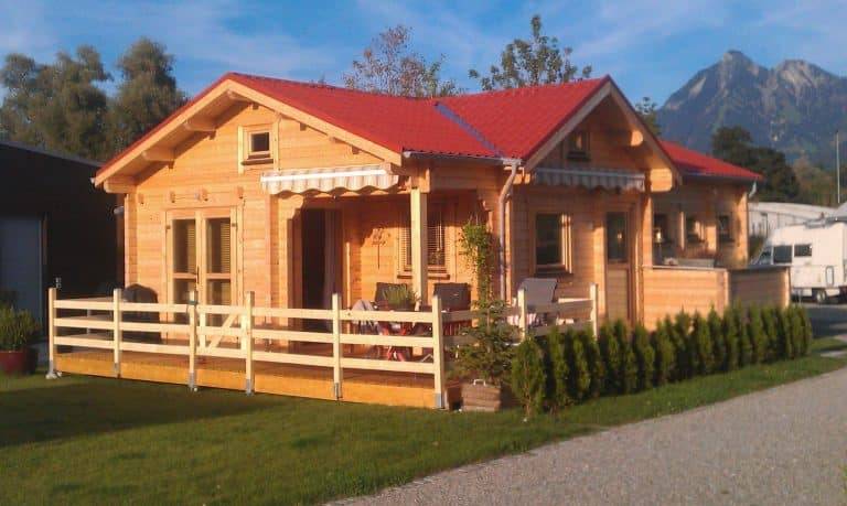 Holzhütte mit rotem Dach und Veranda, gelegen in einer freien Gegend mit Bergen im Hintergrund.