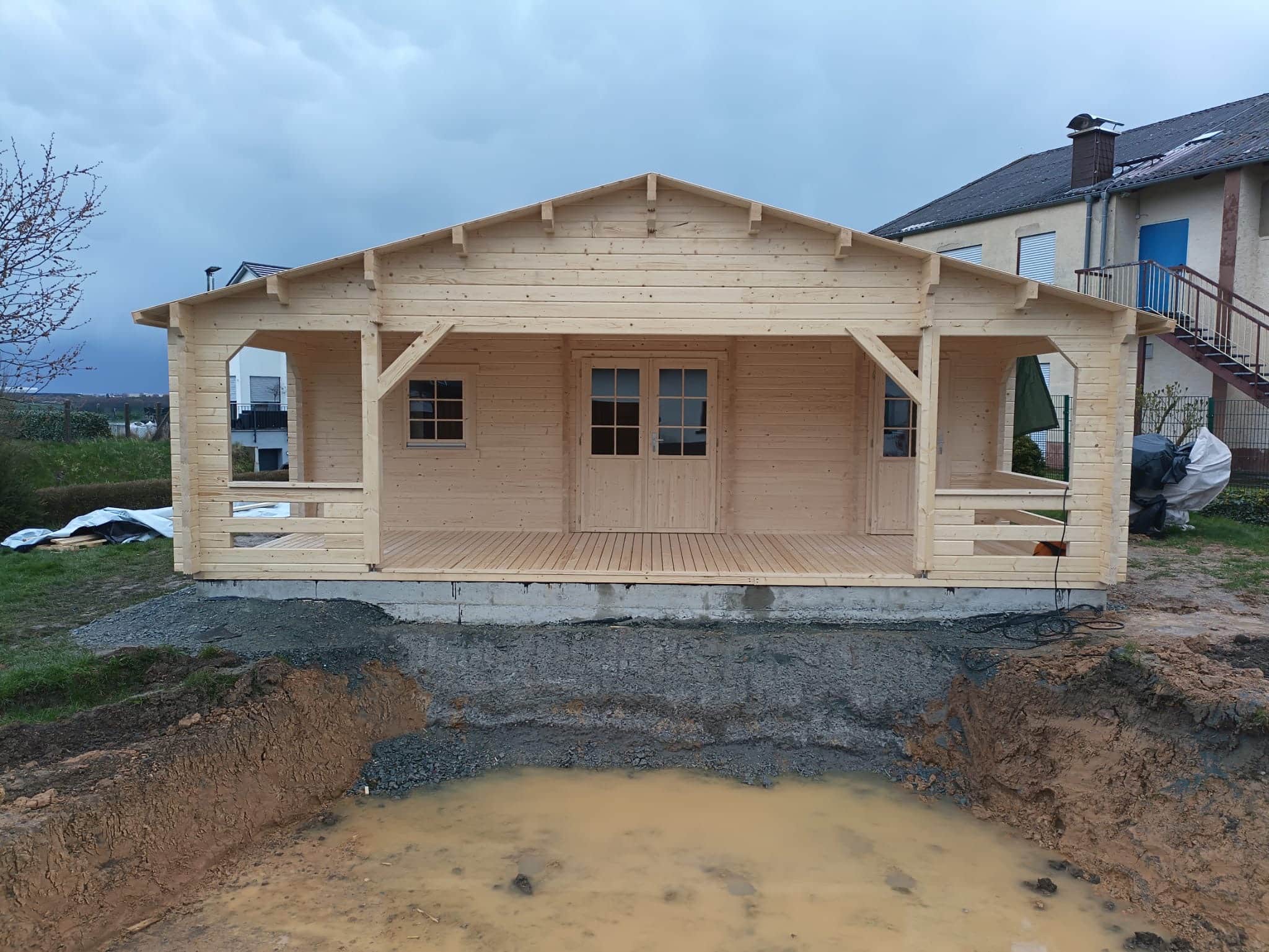 Een nieuw gebouwde houten hut met een overdekte veranda, gelegen op een modderige bouwplaats, onder een bewolkte hemel. De hut, een Tuinhuis Friedland speciale versie, wordt geflankeerd
