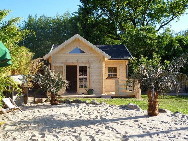 Eine kleine Holzhütte mit Satteldach und Veranda ist von Sand und Palmen umgeben. Unter einem klaren blauen Himmel liegt eine üppig grüne Kulisse aus Bäumen und Pflanzen.