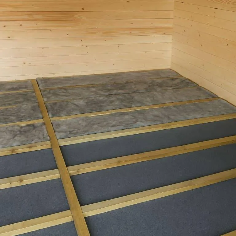 Ein hölzerner Raum mit einem im Bau befindlichen Fußboden; Holzbalken werden über Dämmmaterial verlegt, so dass ein Gerüst entsteht, auf das wahrscheinlich Dielen gelegt werden.